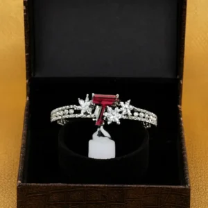 White-gold plated bracelet with a ruby baguette-cut center stone and floral CZ clusters displayed in a black jewellery box by Risha Fine Jewels.